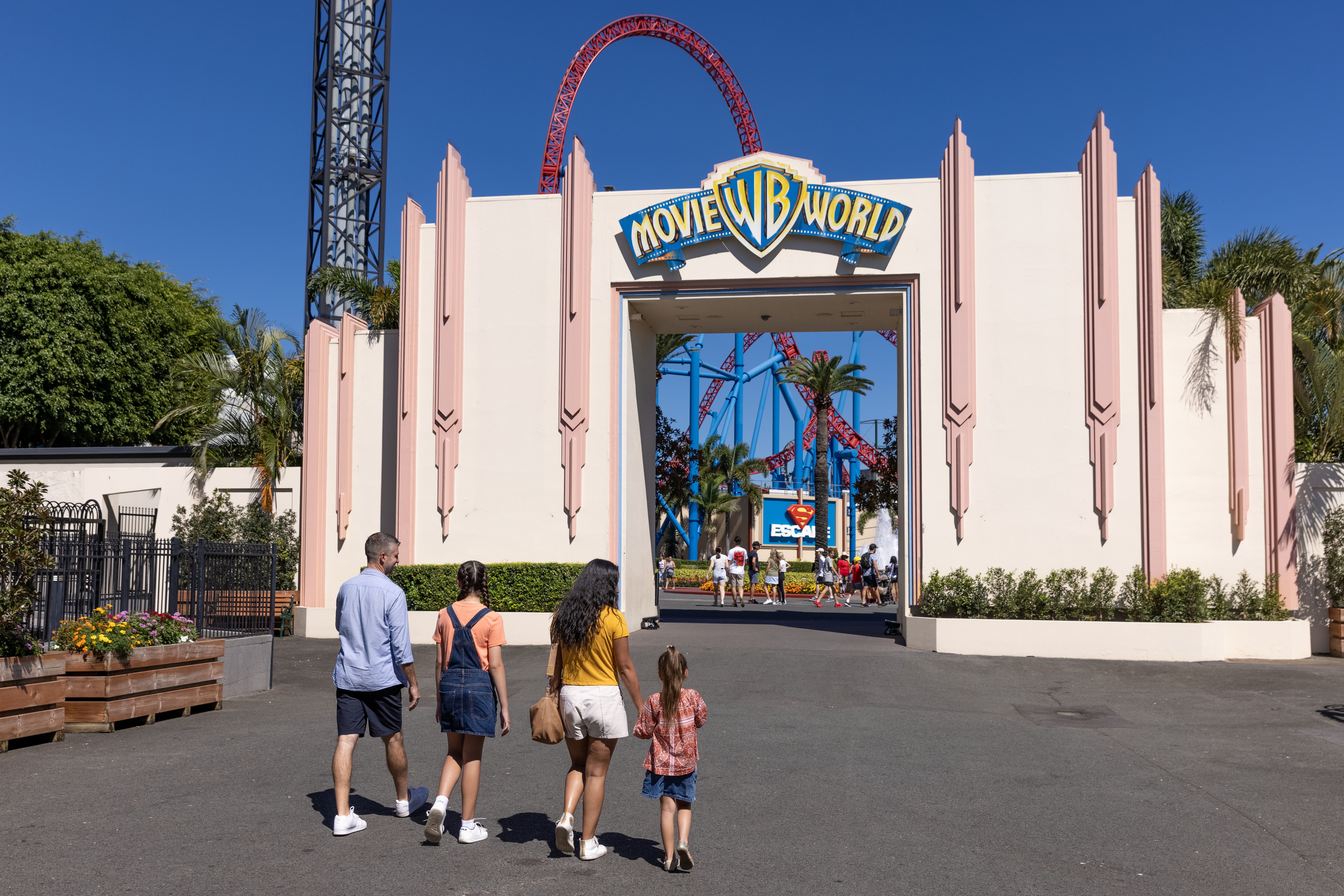 A family of four is approaching the iconic entrance of Movie World on The Gold Coast. A tall red rollercoaster track is located behind it. A family of four is approaching the iconic entrance of Movie World on The Gold Coast. A tall red rollercoaster track is located behind it.
